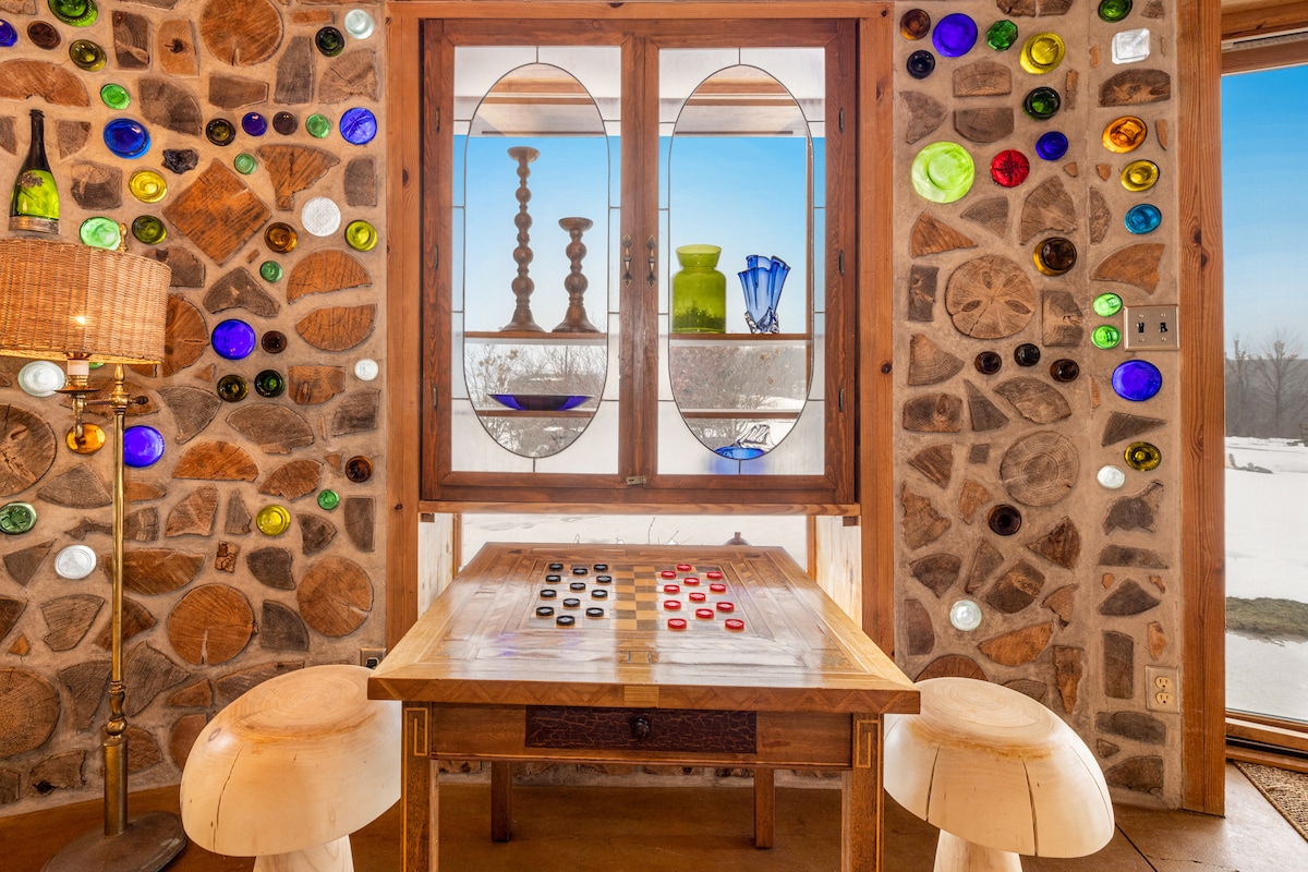 A unique interior space features a table with a checkerboard design, set against a backdrop of natural stone walls adorned with colorful glass decor. The large window offers a view of the outdoor landscape, with decorative vases and candlesticks displayed prominently.