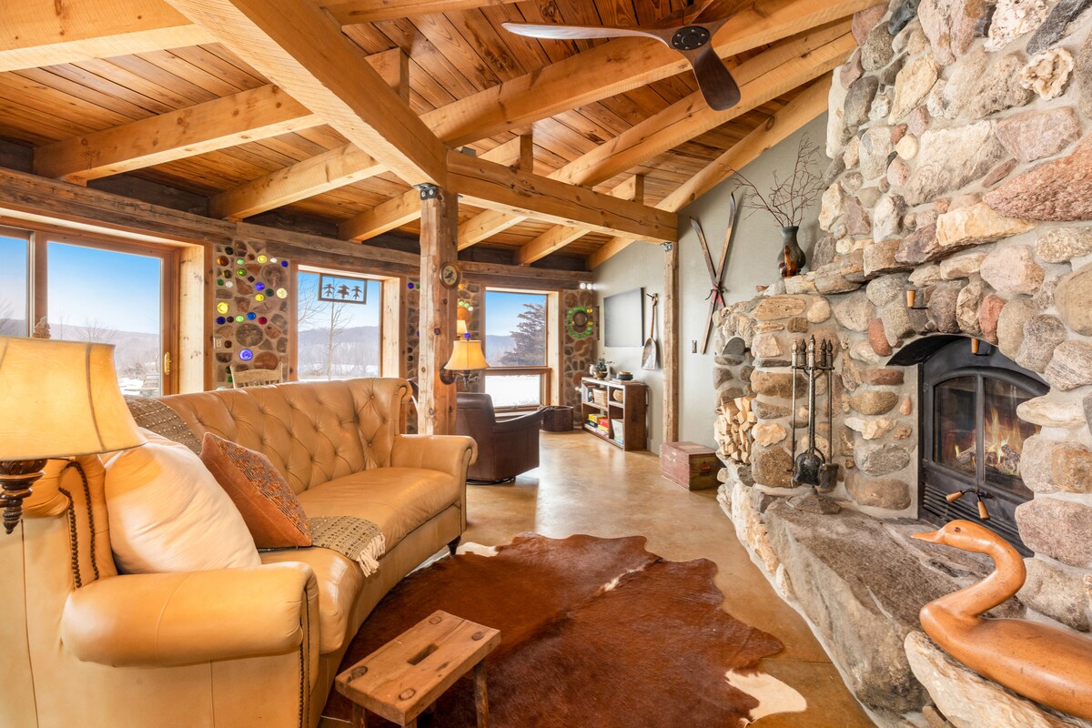 A spacious living area is enhanced by natural wood beams and a stone fireplace, with a comfortable tan sofa and a decorative throw blanket. Large windows provide views of the surrounding landscape, and unique decor elements, such as artwork and a cowhide rug, add character.