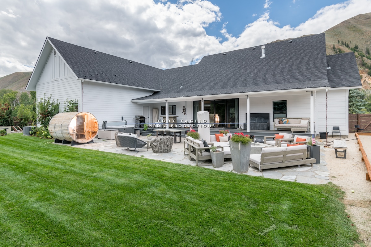 A spacious backyard area is featured, equipped with comfortable seating and a large outdoor dining space. A sleek sauna is positioned nearby. Lush green grass surrounds the patio, complemented by stone pathways and flower planters, with mountains visible in the background under a partly cloudy sky.