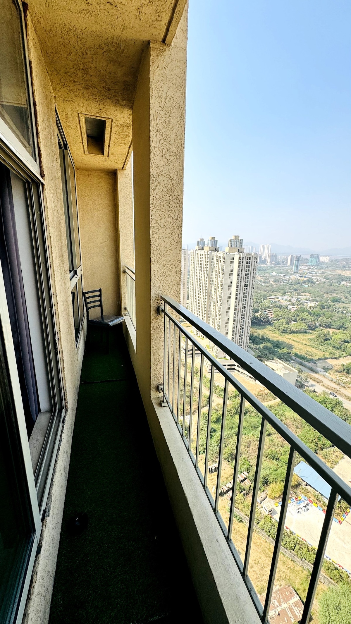 An expansive balcony stretches alongside the apartment, offering unobstructed views of the cityscape below. A single chair is positioned for relaxation, and greenery is visible in the distance, enhancing the scenic backdrop against a clear blue sky.