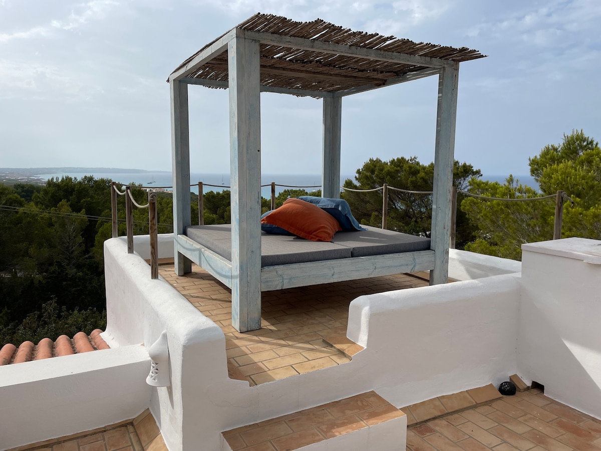 An outdoor lounge area is presented on the rooftop, featuring a shaded wooden structure with a spacious daybed. Brightly colored cushions are arranged comfortably on the bed, and a scenic view of the surrounding landscape and coastline can be seen in the background.