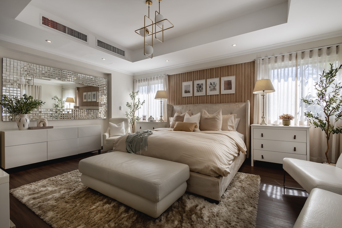 The spacious bedroom features a large bed with soft bedding, complemented by light-toned furniture. Natural light filters through sheer curtains, illuminating the room, which includes decorative plants and a plush area rug. A mirrored wall adds depth, enhancing the elegant design.