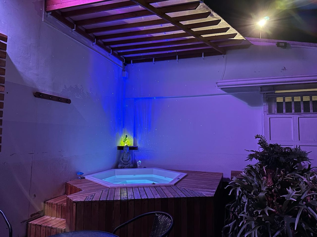 A relaxing outdoor area featuring a wooden hot tub surrounded by plants. Soft lighting casts a soothing glow on the wall, while a small statue adds a serene touch. This space is ideal for gatherings or quiet evenings.