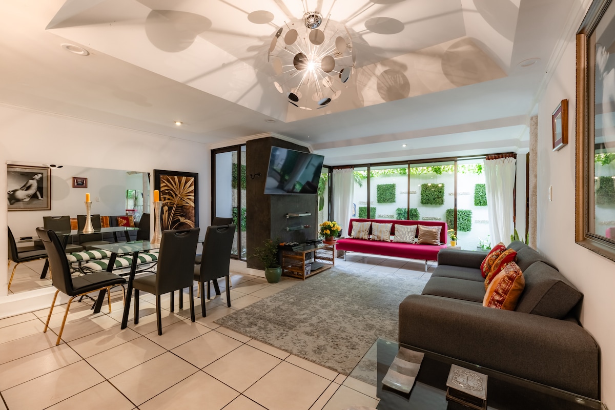 An inviting living area features a modern chandelier overhead and a combination of a comfortable gray sofa and a red accent couch. A glass dining table with black chairs complements the space, while large windows allow natural light to highlight the decorative elements, including wall-mounted greenery.