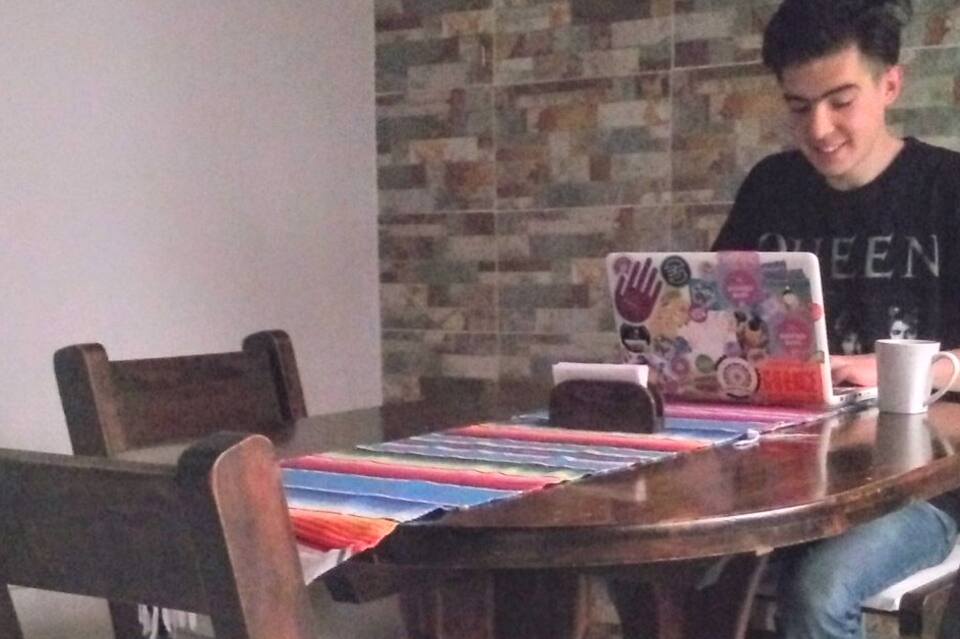 A wooden dining table is set with a colorful striped table runner. A laptop is placed atop the table, accompanied by a small cup. The background features a textured stone wall, adding a neutral tone to the space.