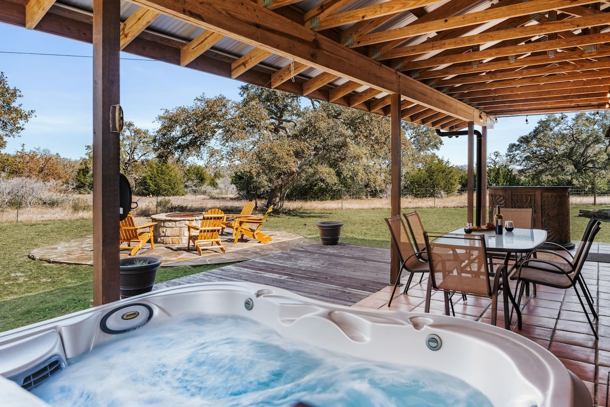 The covered back patio features a hot tub with bubbling water and views of the expansive outdoor space. A fire pit with bright yellow chairs is situated in the distance, surrounded by lush greenery under a clear blue sky.