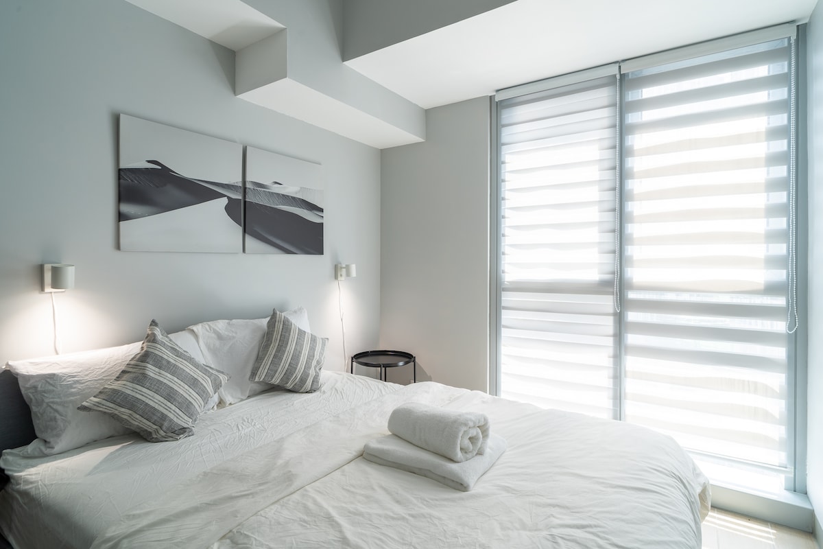 A spacious sleeping area features a large bed dressed in crisp white linens, complemented by striped decorative pillows. Natural light filters through sheer window coverings, illuminating the neutral-toned walls and minimalistic decor, including a bedside table and modern wall sconces.