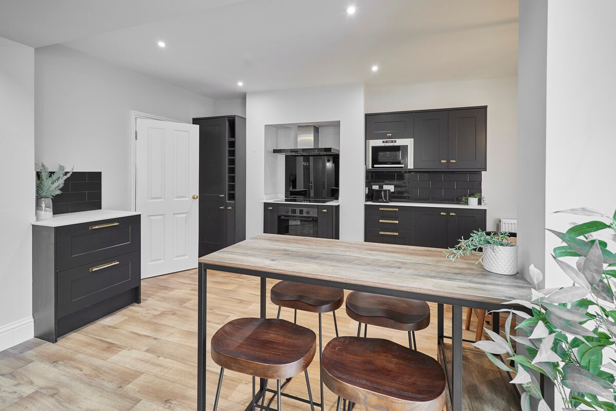 A modern kitchen features sleek black cabinetry and stainless steel appliances, including an electric oven and microwave. A wooden dining table with four stools is situated in the foreground, while a door leads to another room. Indoor plants add a touch of freshness to the space.