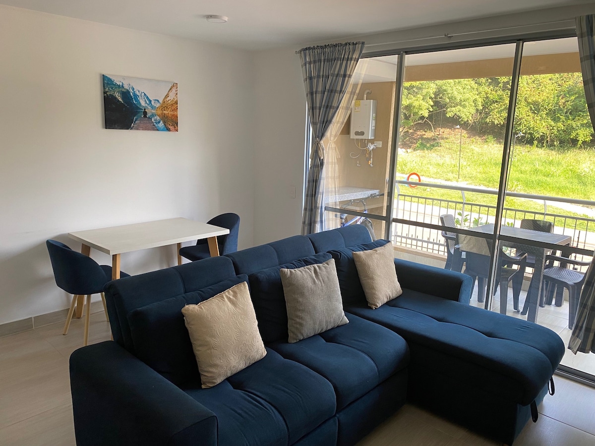 A modern living area features a dark blue sectional sofa with pillows, positioned adjacent to a dining table suited for four. Large sliding glass doors provide access to a balcony, allowing natural light to illuminate the space. A framed landscape photograph adds visual interest to the wall.