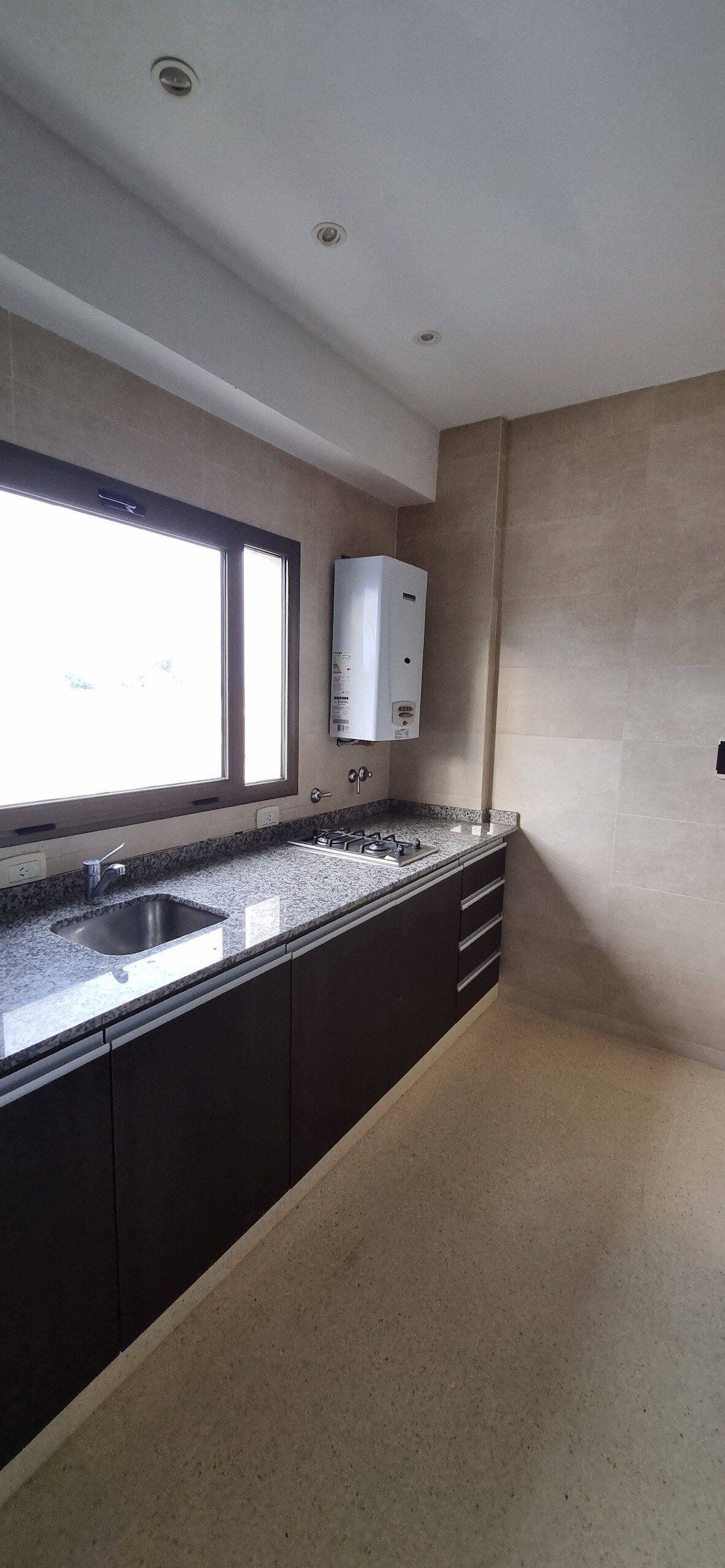 A modern kitchen area is presented, featuring dark cabinetry against light stone walls. A window allows natural light to enter, illuminating a countertop equipped with a sink and kitchen appliances. The floor is smoothly finished, enhancing the spacious feel of the area.