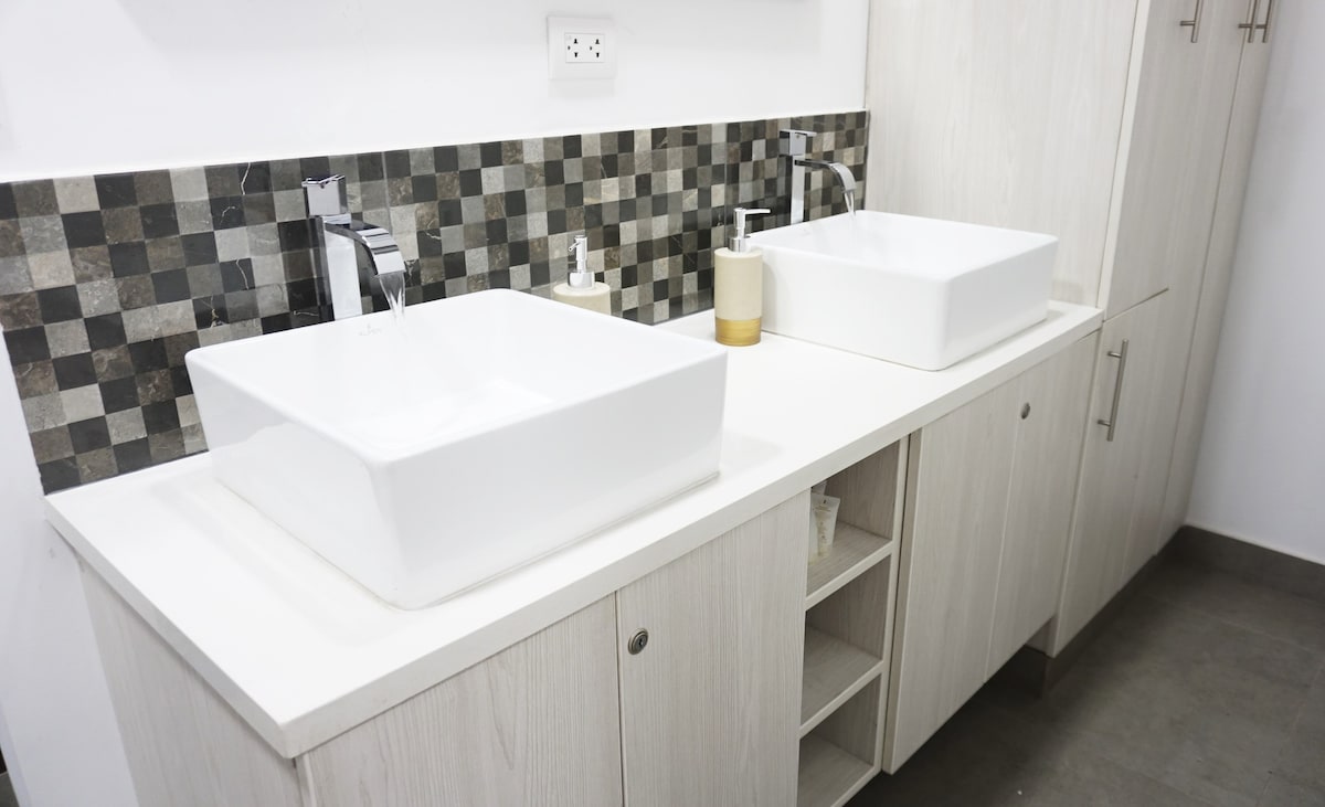 A modern bathroom features dual white square sinks positioned on a light countertop. A mosaic of textured gray and beige tiles creates a striking backdrop. Underneath, cabinetry provides additional storage, enhancing the room's functionality.
