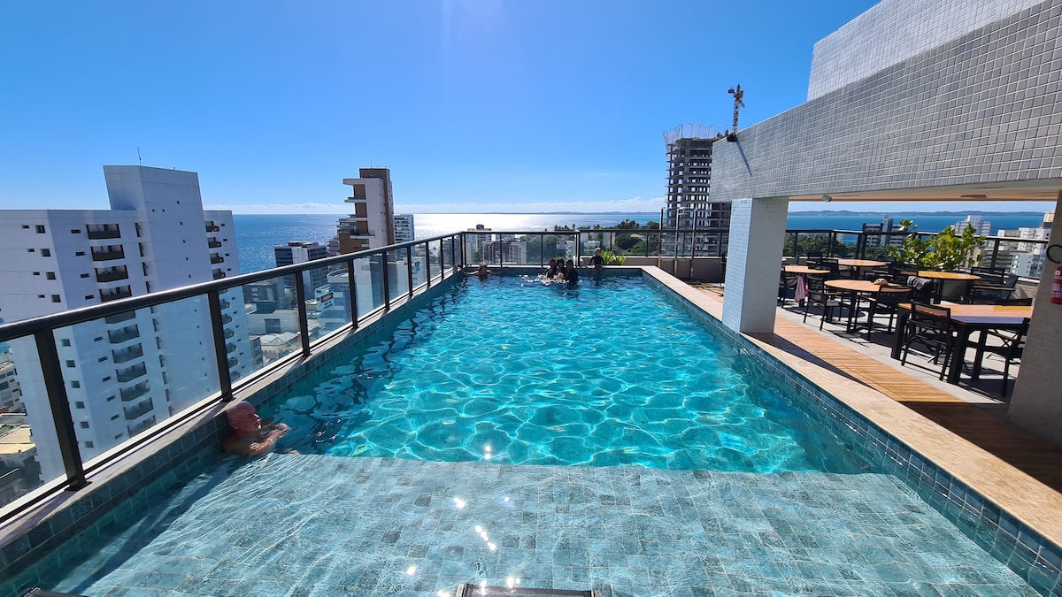 A rooftop pool is visible, showcasing clear turquoise water surrounded by a glass railing. Sun loungers and tables are positioned near the pool, with a panoramic view of the skyline and ocean in the background, under a bright blue sky.