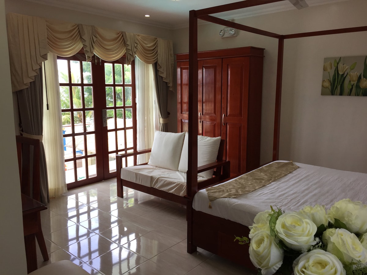 A bright room features a four-poster bed and a seating area with two wooden chairs. Light drapes frame the glass doors that open to a balcony. A wooden wardrobe stands against the wall, and a bouquet of white flowers adds a touch of softness.