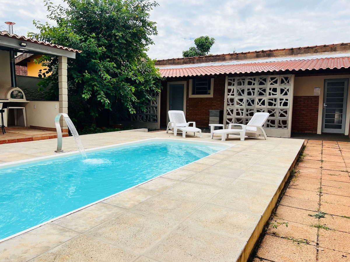 A swimming pool is centered in the outdoor area, featuring a gentle waterfall and surrounded by a spacious deck. Two white lounge chairs are placed nearby, with greenery enhancing the environment. A covered patio area can be seen in the background, offering a cozy gathering space.