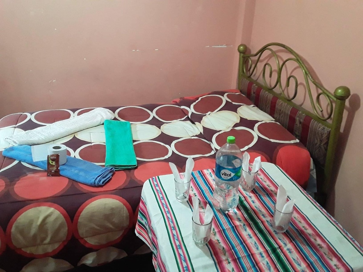 A cozy room featuring a small bed adorned with patterned linens. A table is set with water bottles and glasses, while neatly folded towels are placed on the bed and table. The walls are painted in a soft pink hue, contributing to a welcoming ambiance.