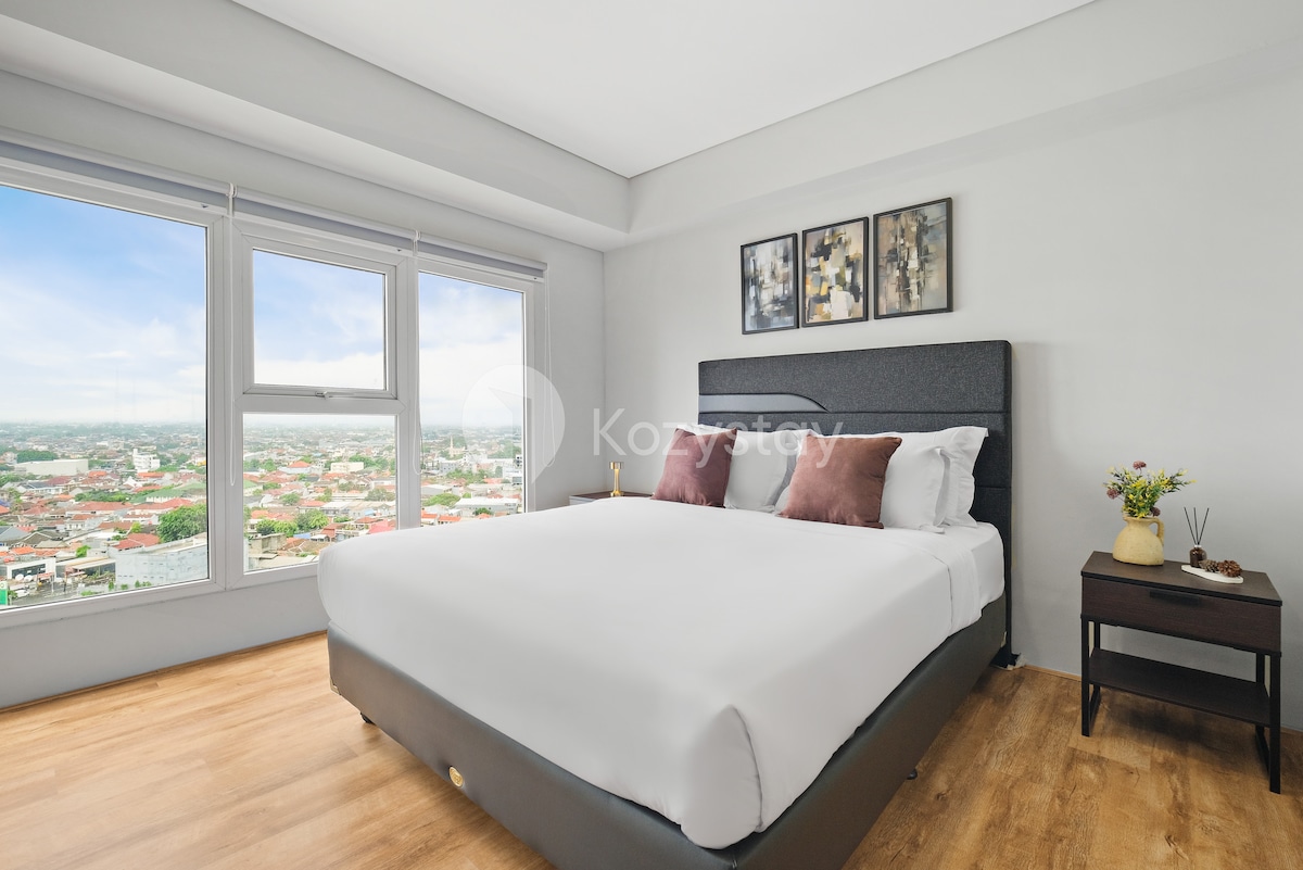 A well-furnished bedroom features a spacious bed with crisp white linens and two decorative pillows. Natural light floods in through large windows showcasing sweeping urban views. Artwork adorns the wall above the bed, complemented by a small nightstand holding decorative elements.