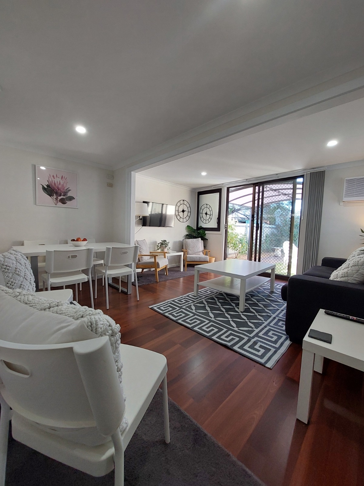 A bright living area features a comfortable seating arrangement with a dark sofa and armchairs, complemented by a light coffee table. Natural light floods in through sliding glass doors, leading to an outdoor space. A dining table for six is positioned nearby, enhancing the open layout.