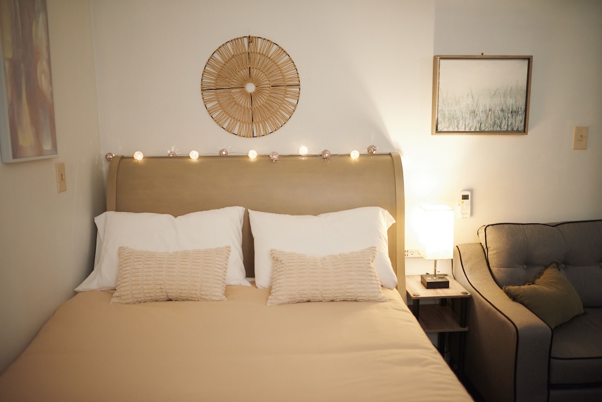 A cozy sleeping area features a comfortable bed with soft, neutral linens and decorative pillows. Fairy lights are placed above the headboard, subtly illuminating the space. A small lamp and a framed piece of artwork are positioned beside a full couch.