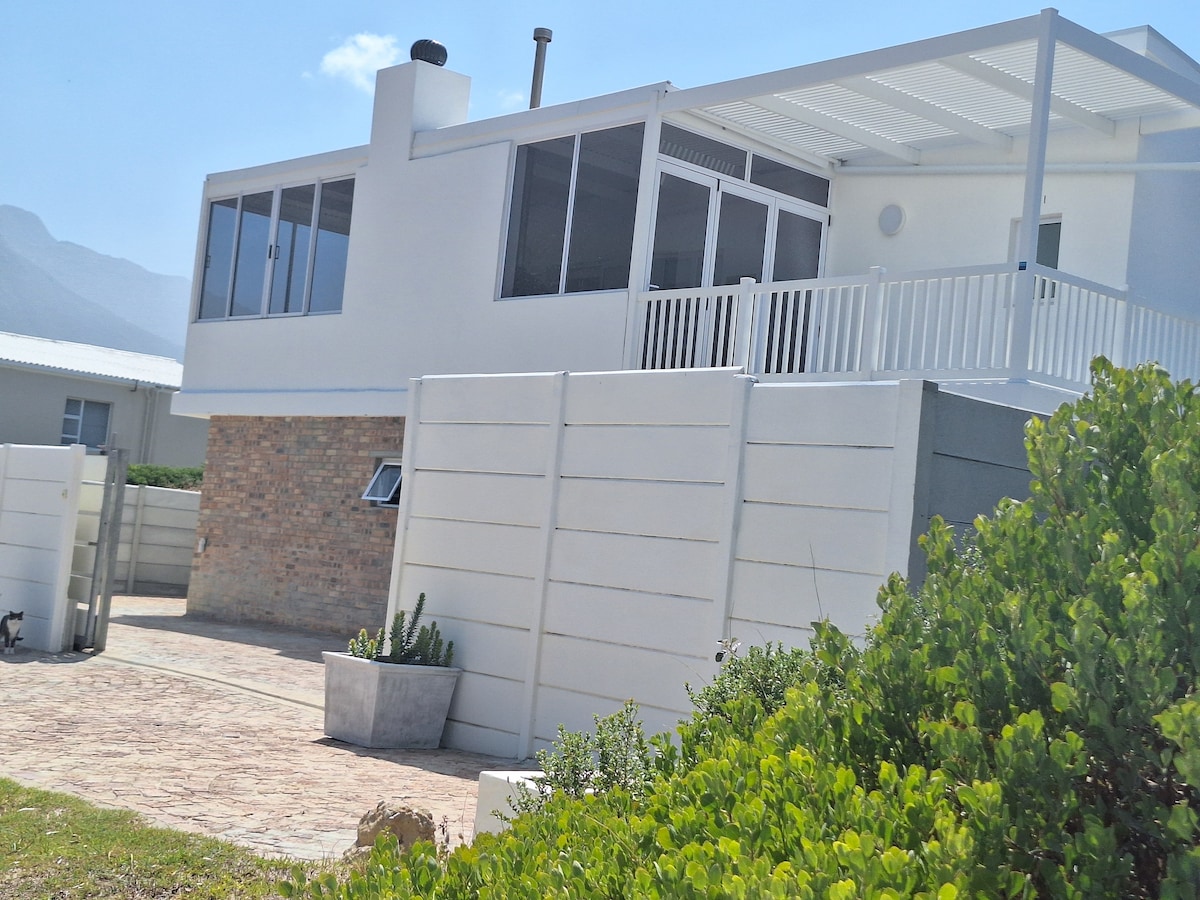 A modern two-story building with a white facade is framed by lush greenery. Large windows reflect sunlight, while an elevated terrace offers outdoor seating. A stone pathway leads to a secure entrance, ensuring privacy and a welcoming ambiance.