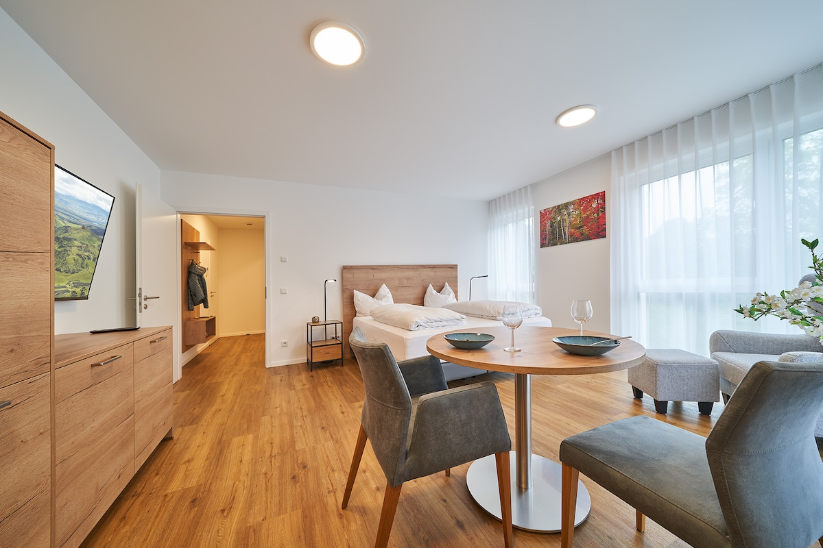 An open living-sleeping area is highlighted by a spacious bed and a circular dining table set for two. Soft lighting illuminates the wood-paneled furniture, while large windows allow natural light to fill the room. A painting adds a touch of color to the neutral decor.