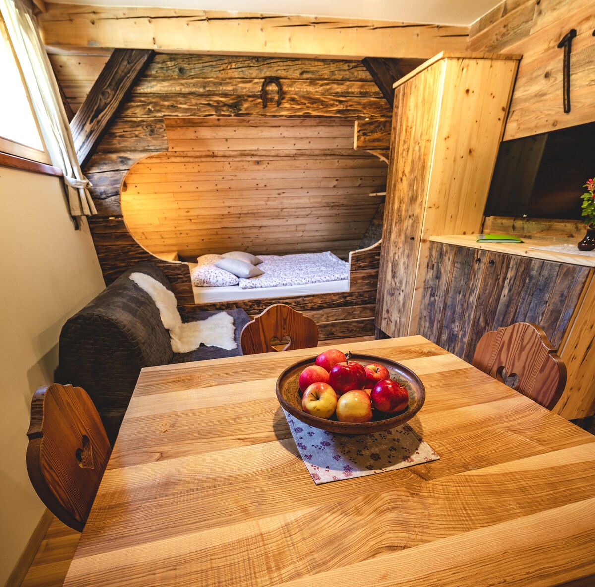 A wooden dining table is centered in the space, surrounded by carved chairs. A bowl of red and green apples rests on the table. The room features rustic wood paneling and a cozy sleeping nook with a comfortable bed, softly lit by natural light.
