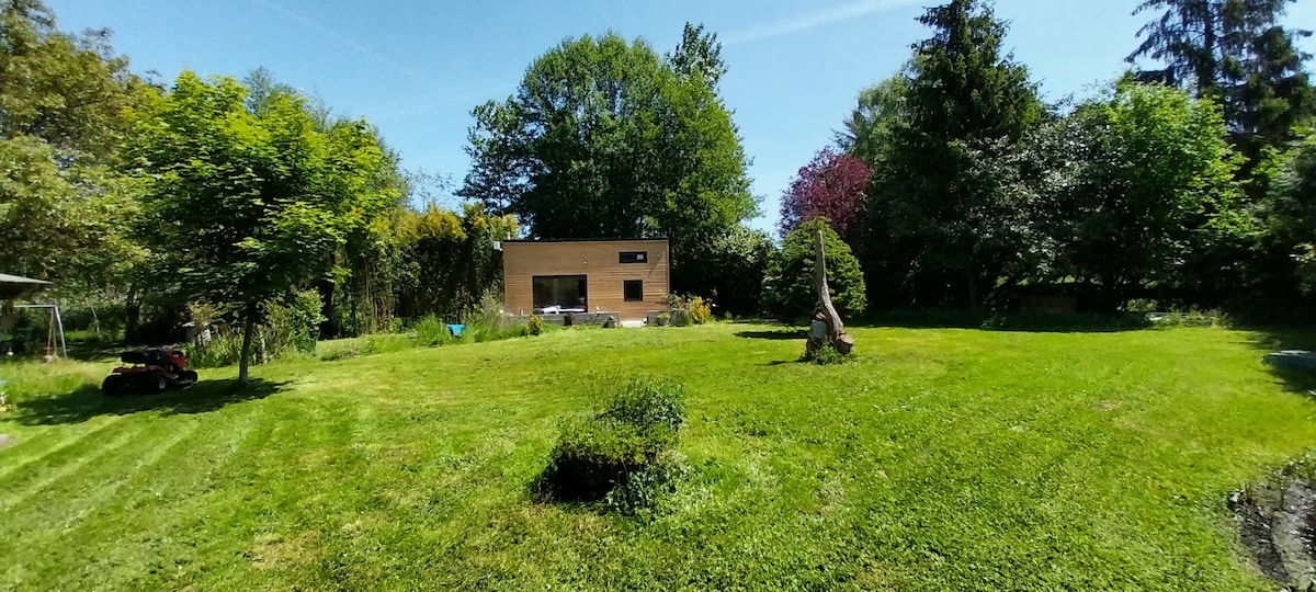 A spacious garden area is surrounded by lush greenery and trees. The well-maintained lawn features manicured grass and varied plant life, creating an inviting outdoor space. Sunlight filters through the trees, offering a serene ambiance, which can be enjoyed from the terrace.