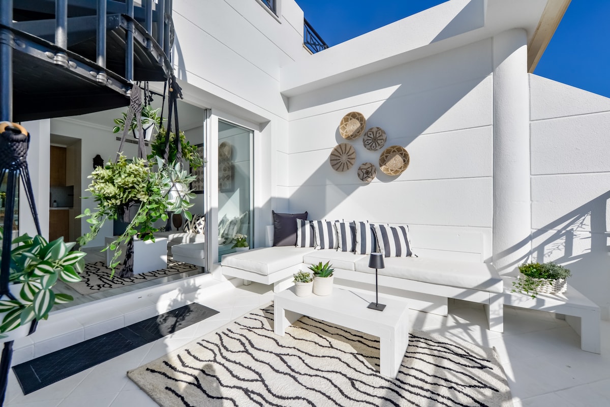 An outdoor seating area features a white sectional sofa adorned with striped cushions. A small black lamp sits on a white coffee table, surrounded by potted plants. Textured rugs enhance the space, while decorative wall plates add visual interest to the bright, airy environment.