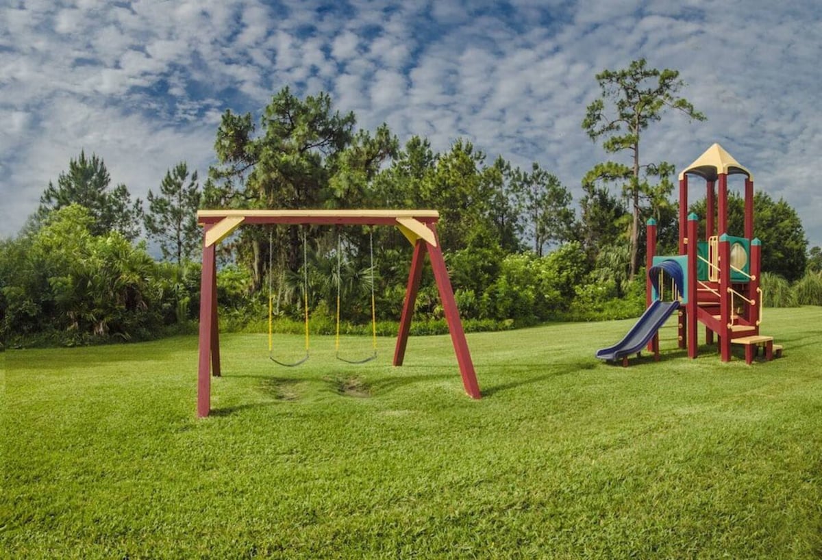 An outdoor play area is presented, featuring a vibrant red swing set and a colorful play structure with a slide. Lush green grass surrounds the playground, complemented by trees in the background under a partly cloudy sky.