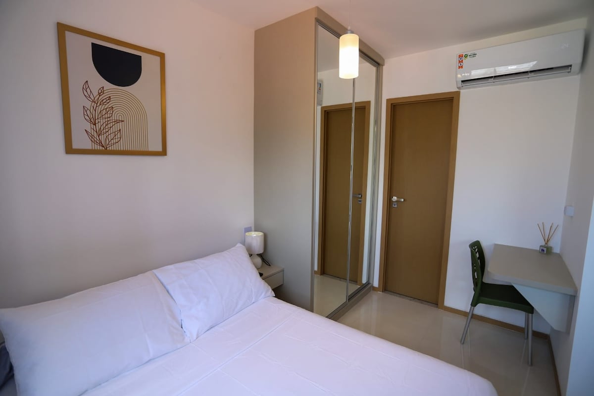 A well-lit bedroom features a spacious bed with crisp white linens. A wall-mounted artwork adds a subtle design touch. A large mirror is positioned on the wardrobe, reflecting the space. A small workspace with a chair is located near the door, enhancing functionality.