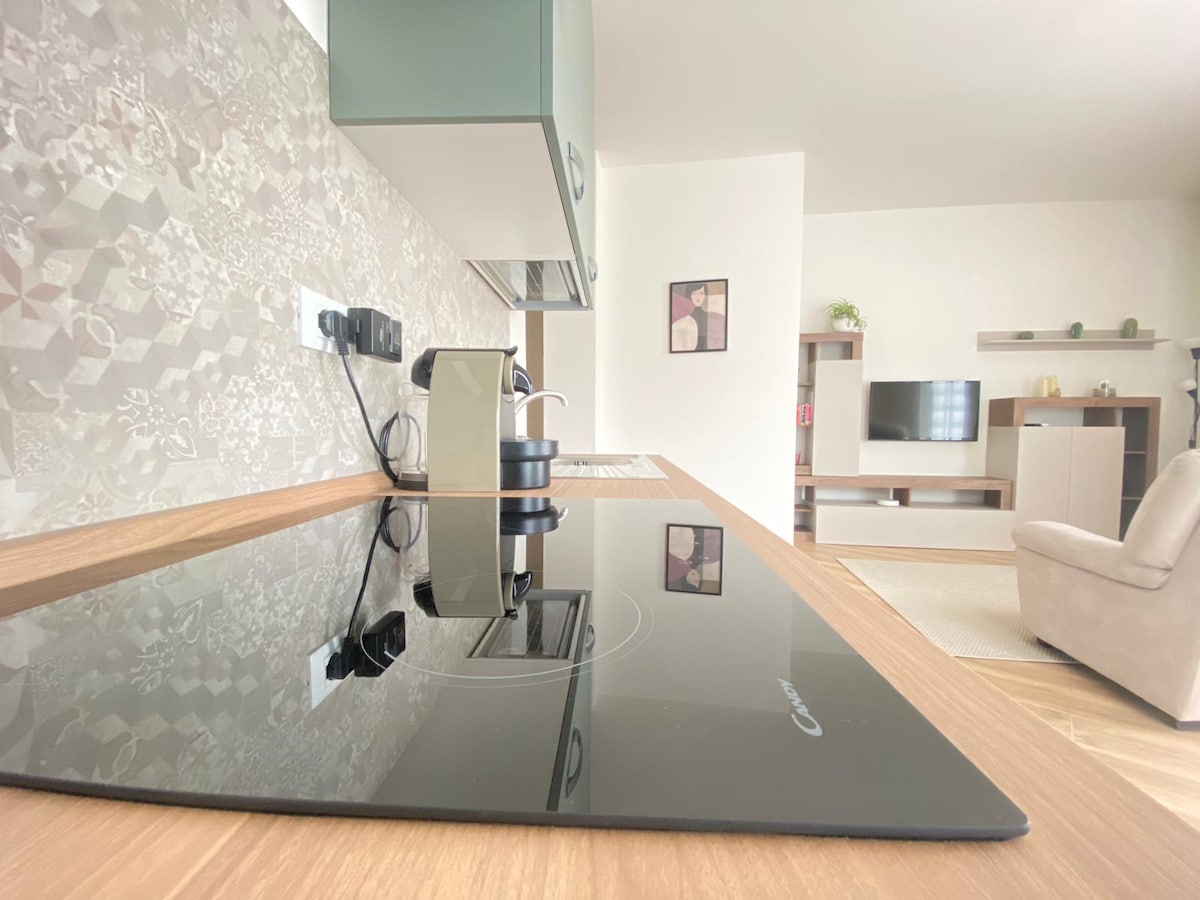 A modern kitchen area features a sleek induction cooktop and a minimalist coffee maker, complemented by stylish cabinetry. The backsplash showcases a patterned design, while the adjoining living space is visible in the background, highlighted by a light-toned accent wall and contemporary furniture.
