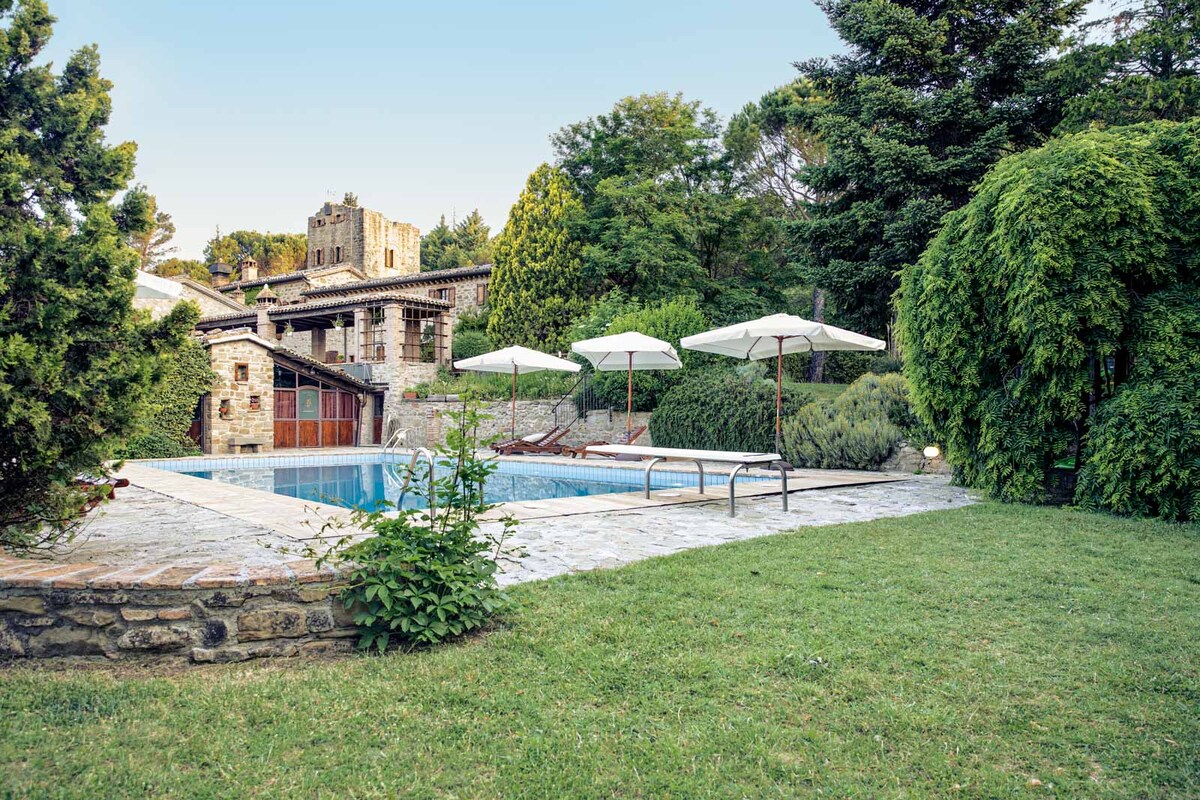 A serene outdoor area features a swimming pool surrounded by stone paving and lush greenery. Two umbrellas provide shade over sun loungers, inviting relaxation. The property is framed by trees and a well-maintained lawn, showcasing a charming rustic building in the background.