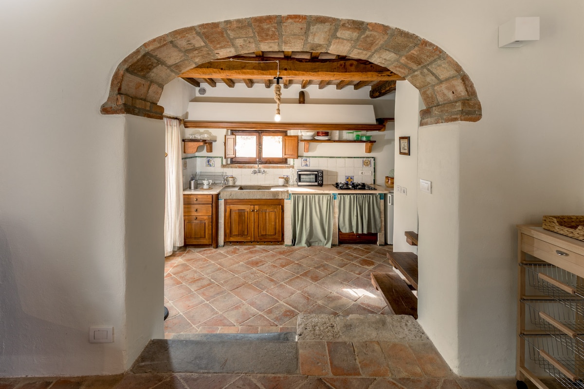 The kitchen area features exposed wooden beams and terracotta tile flooring. A wooden table is accompanied by a classic cooking stove, with a window allowing natural light. Cabinets with a rustic finish complement the space, providing a cozy and functional atmosphere.