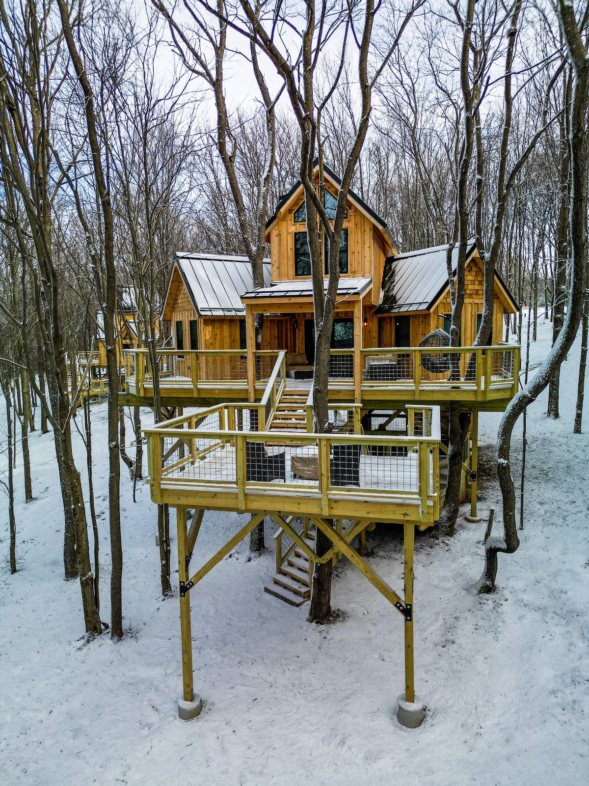 A custom-built treehouse sits elevated among leafless trees, surrounded by a blanket of snow. Its wooden exterior complements the natural surroundings, while expansive decks provide outdoor space. Stairs lead to the entrance, inviting guests to explore the unique retreat above the forest floor.