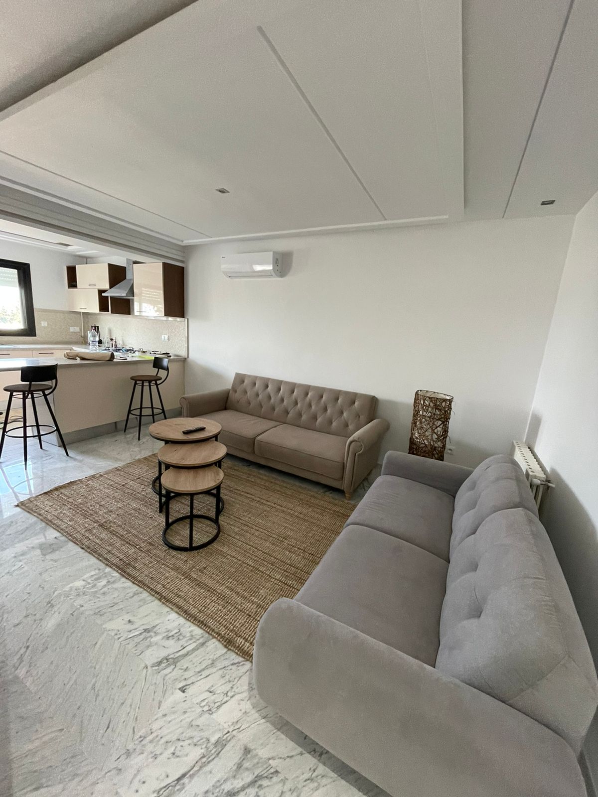 A modern living area is presented with a light-colored, two-seater couch and a three-seater couch, arranged around a neutral rug. Two small tables provide surface space. The open kitchen is visible in the background, with bar stools positioned at the counter.