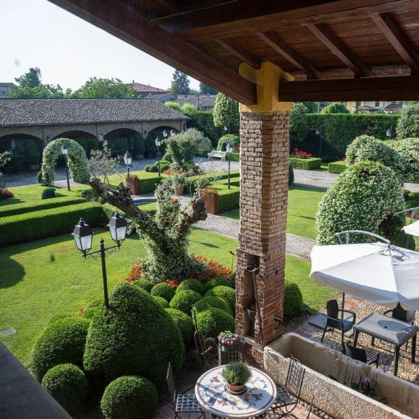 A well-maintained garden features neatly trimmed hedges, vibrant flowers, and a variety of trees. Stone pathways wind through the greenery, leading to seating areas with tables and umbrellas. The space is enhanced by decorative lanterns and a rustic brick structure that complements the natural surroundings.
