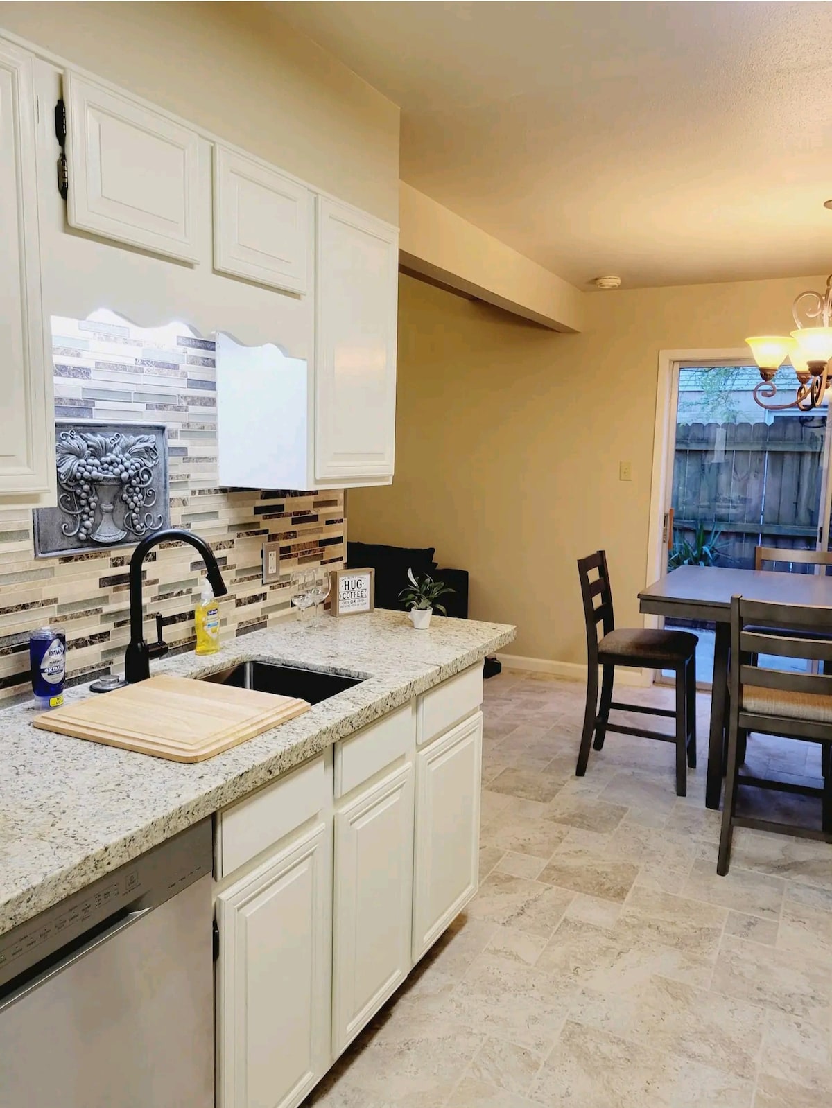 A well-appointed kitchen features white cabinetry and a granite countertop with a cutting board positioned beside a stainless steel sink. A modern backsplash adds texture, while a dining area with a wooden table and two high chairs is visible by a window leading to the outdoor space.