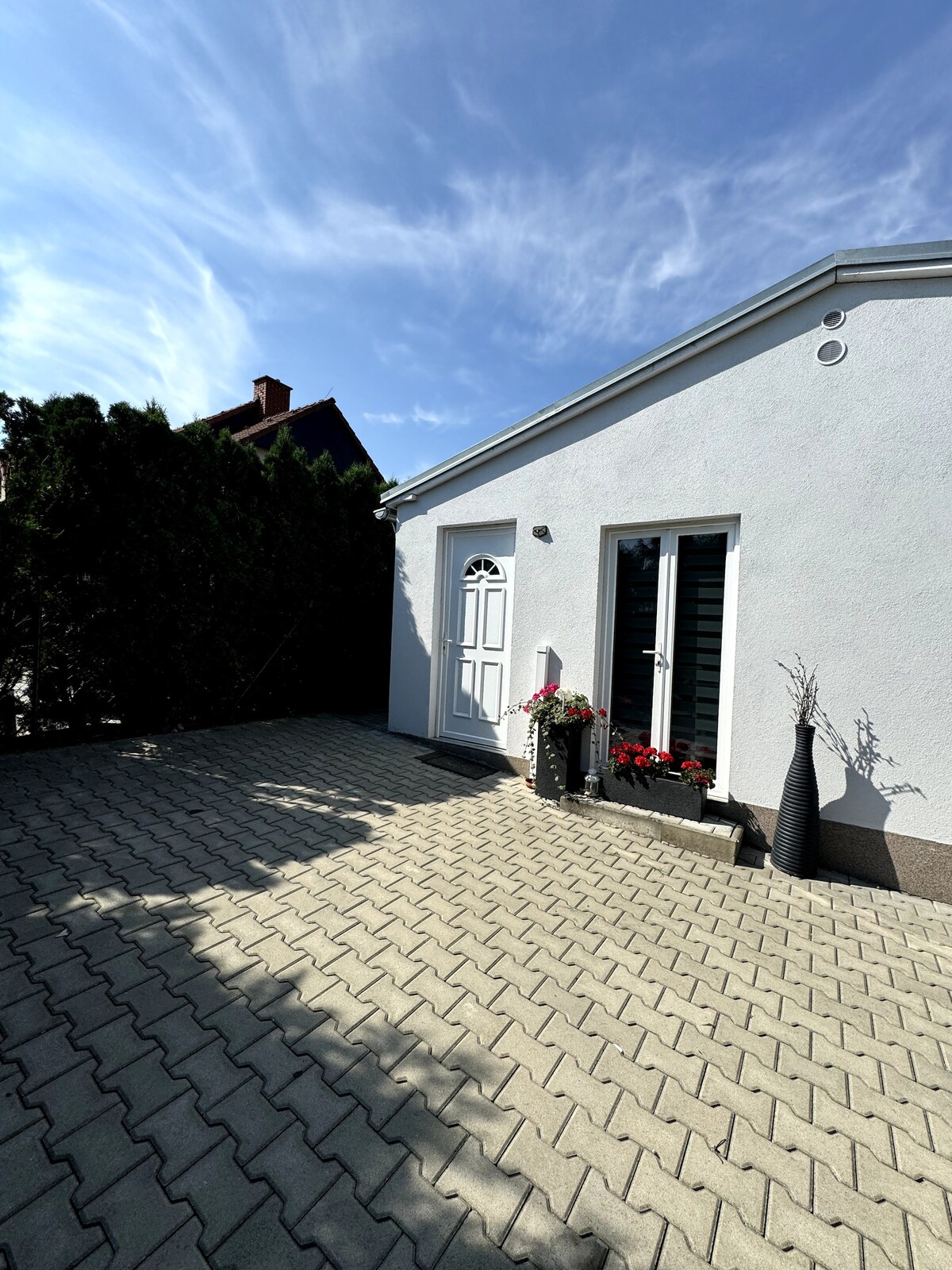 A white structure with a charming door is set against a blue sky. A paved area is featured, bordered by greenery and flower pots. The patio is spacious, inviting outdoor relaxation.