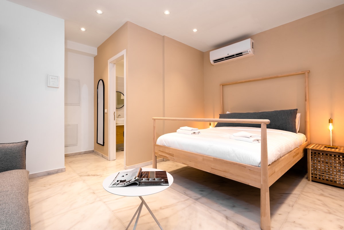 A spacious bedroom features a wooden bed with clean white linens. An inviting sofa sits beside a small table. Warm lighting illuminates the area, complemented by a subtle color palette. A doorway leads to an adjoining bathroom, enhancing the room's accessibility.