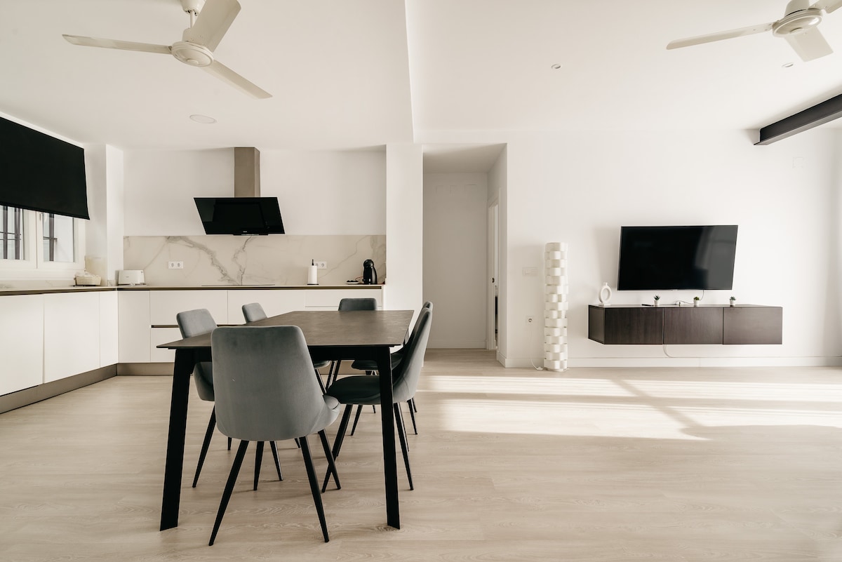 An open-plan dining area features a large table surrounded by six chairs, illuminated by natural light from nearby windows. The adjoining kitchen is designed with modern cabinetry and equipped with essential appliances. A wall-mounted television adds convenience to the space.