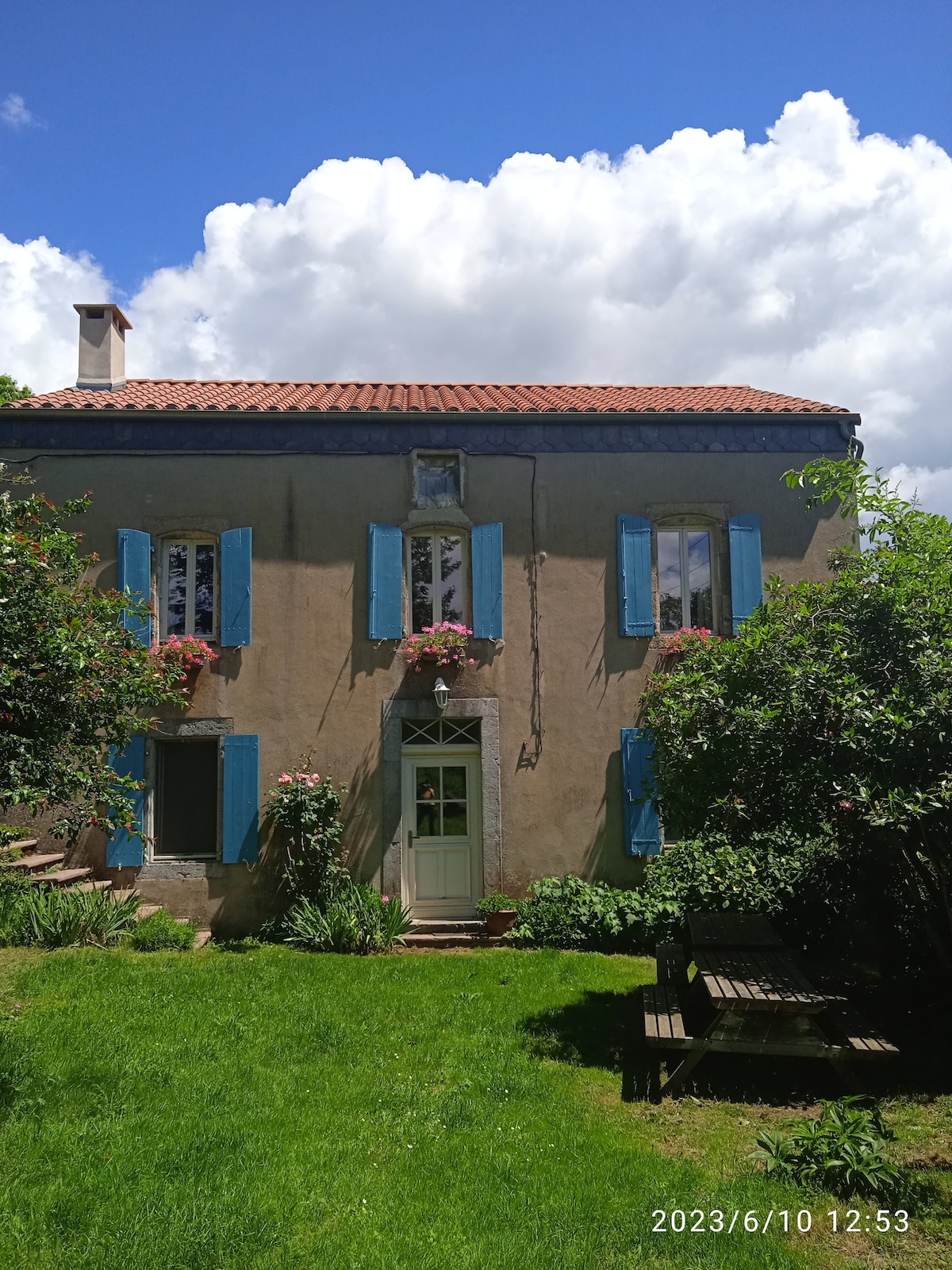 The charming exterior of a two-story house is visible, featuring light-colored walls and blue shutters. Flowers are arranged in window boxes under the windows. A green garden surrounds the house, complemented by a wooden bench. Fluffy clouds float in a bright blue sky above.