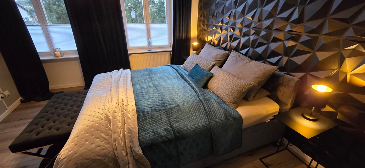 A spacious double room is showcased, featuring a textured black and royal blue accent wall. Soft, ambient lighting is provided by two bedside lamps. A light-colored bedspread covers the spacious bed, accented by coordinating cushions. A bench and window with dark curtains complete the inviting space.