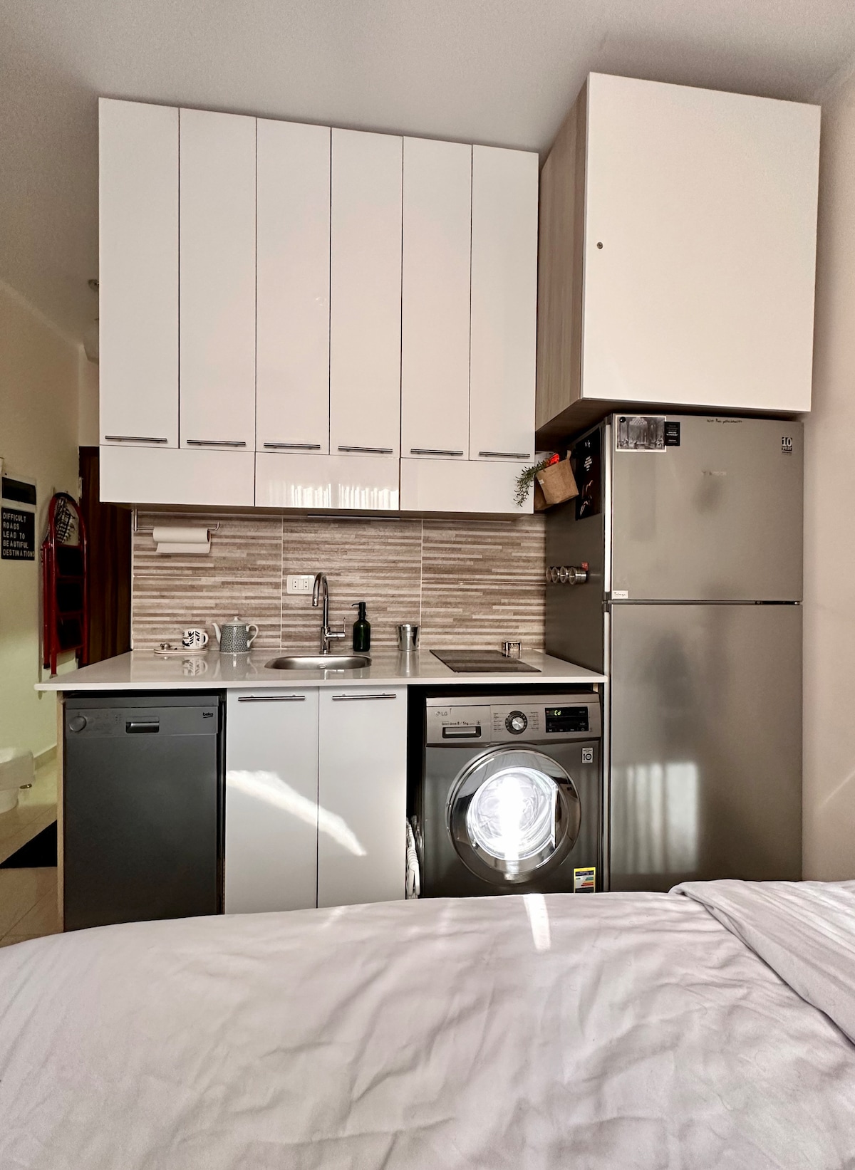 A compact kitchen area is equipped with a refrigerator, washing machine, and dishwasher beneath a wall of sleek storage cabinets. Counter space is available for meal preparation, featuring a sink and essential appliances against a backdrop of textured tiles.