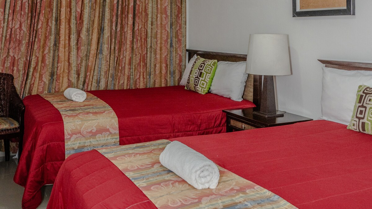 The room features two comfortable beds covered with vibrant red covers, complemented by decorative pillows. A nightstand is positioned between the beds, and a lamp is present for additional lighting. Colorful curtains frame a window, adding a lively touch to the space.