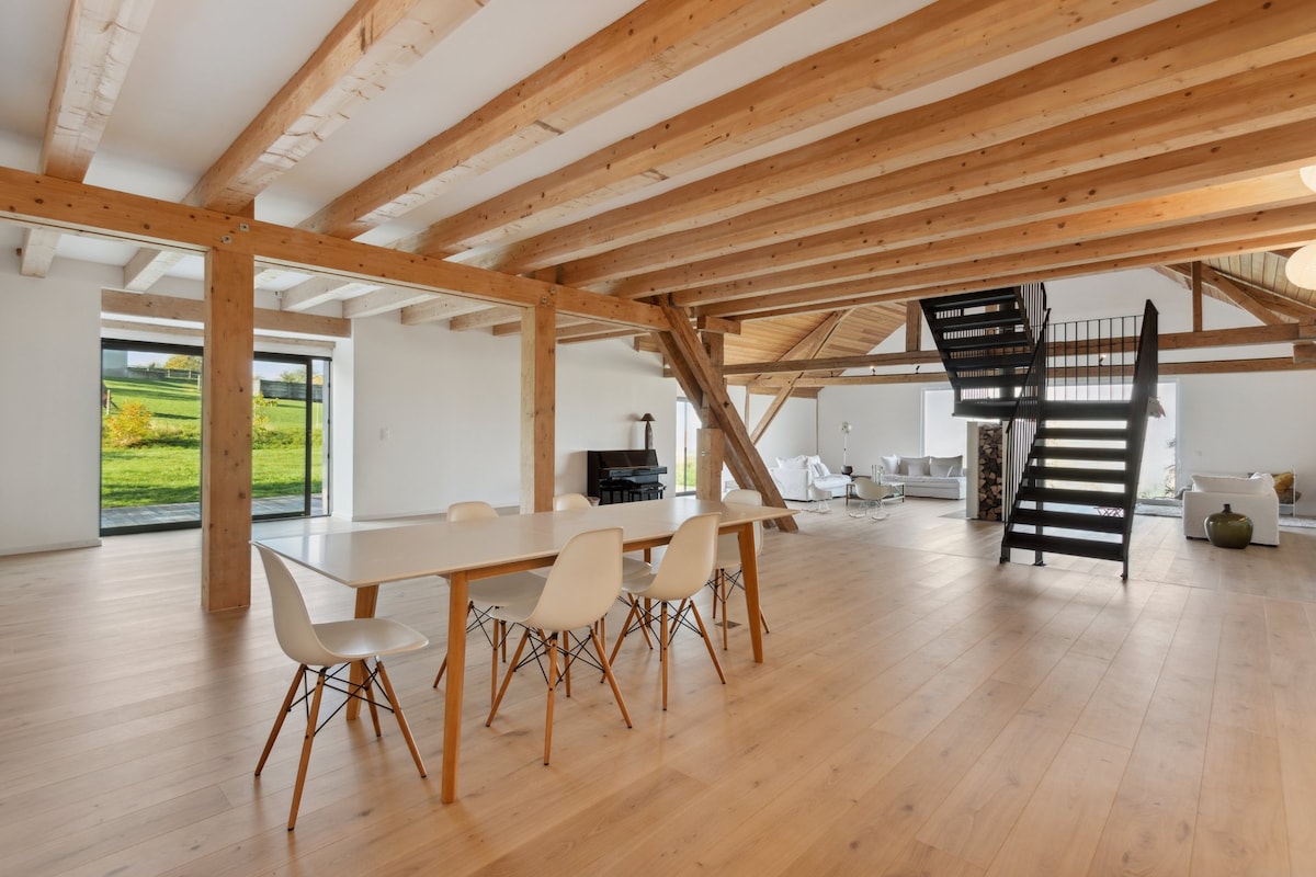 An open living space features a large dining table surrounded by white chairs. Exposed wooden beams enhance the loft's architectural design, while large windows provide views of the greenery outside. A modern staircase leads to an upper level, adding depth to the area.