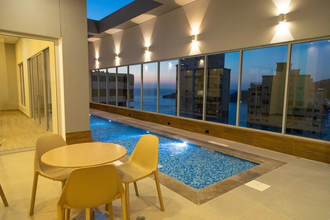 An outdoor area features a small inviting pool surrounded by modern lounge chairs and a circular table with four chairs. Large glass windows provide an unobstructed view of the ocean and city, allowing natural light to illuminate the space.