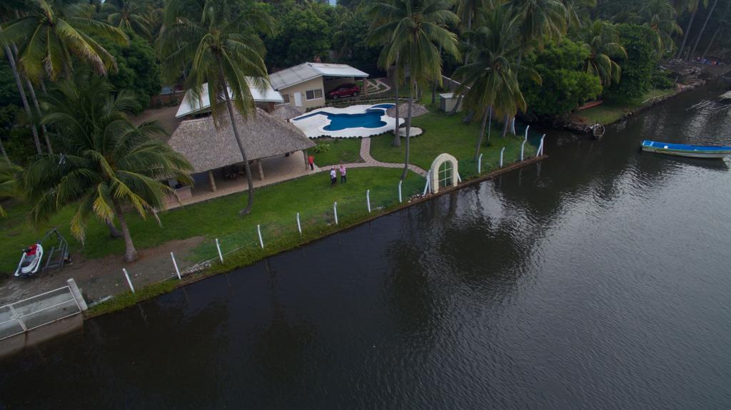 A spacious outdoor area is highlighted by a swimming pool surrounded by lush greenery and palm trees. The property is adjacent to a calm body of water, with a pathway lined by fencing leading along the water's edge.