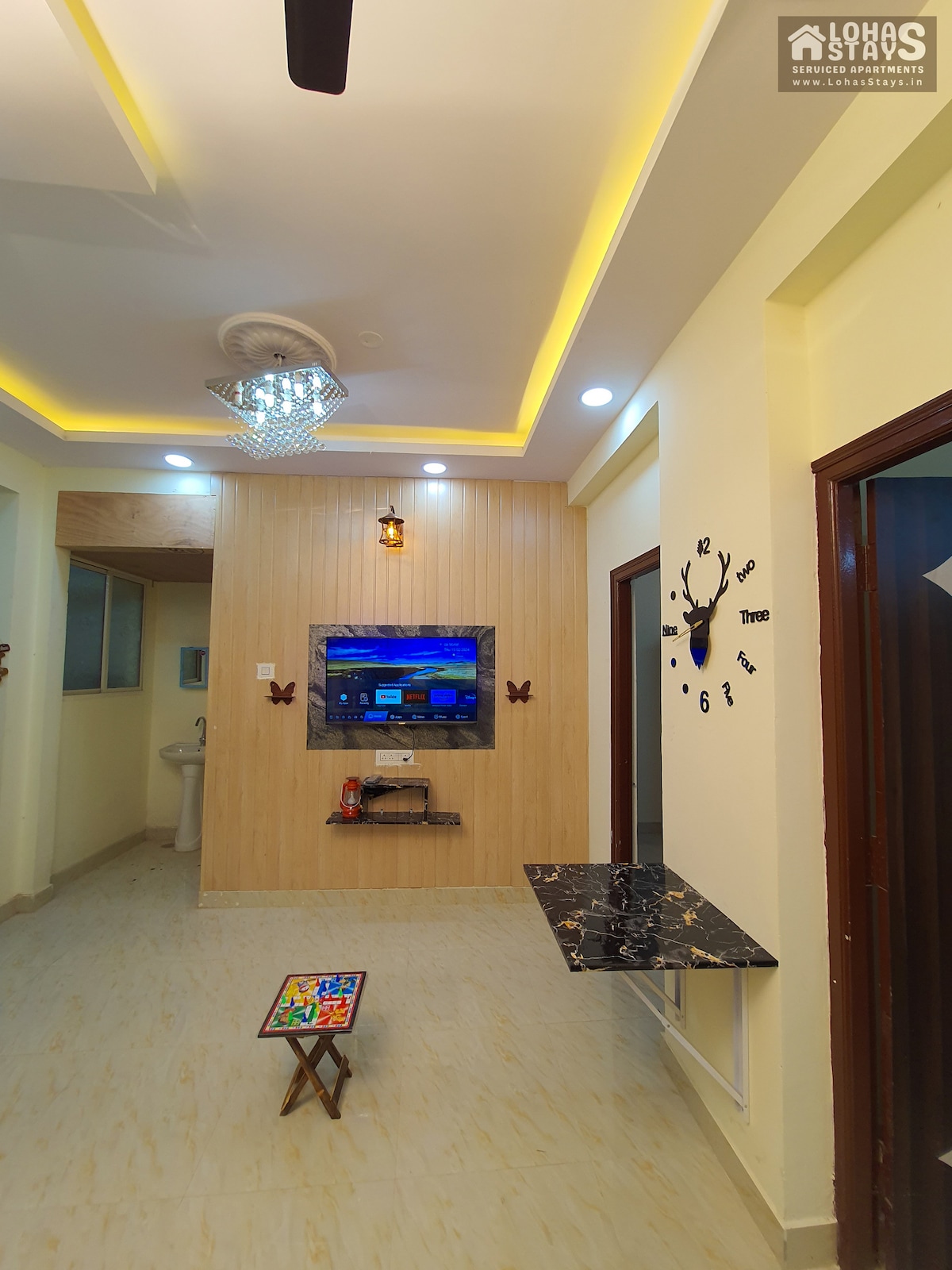 A well-lit living area features modern decor with a wall-mounted 50-inch TV, providing entertainment options. A small, patterned table is positioned in front of the TV, while floating shelves and wall art add visual interest. The overall space exudes a cozy and inviting ambiance.