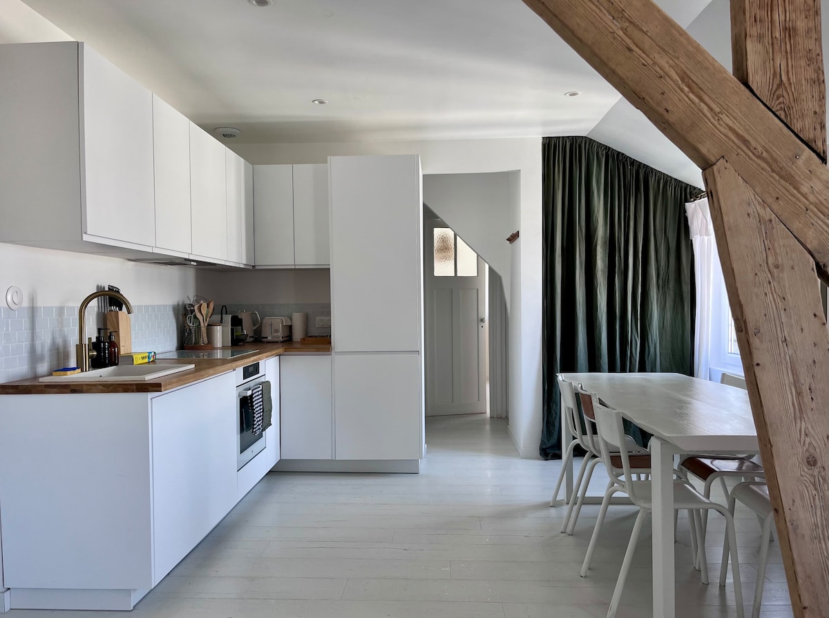 The kitchen features modern white cabinetry and a wooden countertop, equipped with essential appliances including an induction stove, dishwasher, and refrigerator. Natural light pours in through large windows adorned with green curtains, while a simple wooden dining table and chairs are positioned nearby.