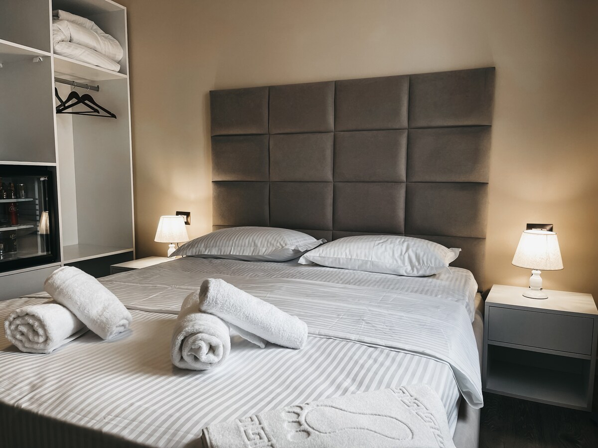A neatly arranged sleeping area features a spacious bed with soft linens and two pillows. Fresh towels are neatly rolled and placed on the bed. Bedside lamps provide soft lighting, while a minimalist wardrobe and a small refrigerator are visible in the background.