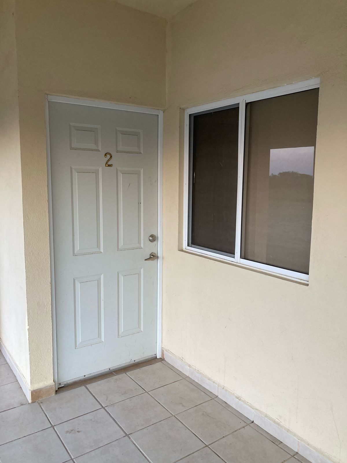 A cream-colored exterior features a simple door numbered '2' aligned with a sliding window nearby. Light-colored tiles cover the floor, and a neutral wall provides a minimalistic backdrop. Natural light filters through the window, illuminating the entryway.